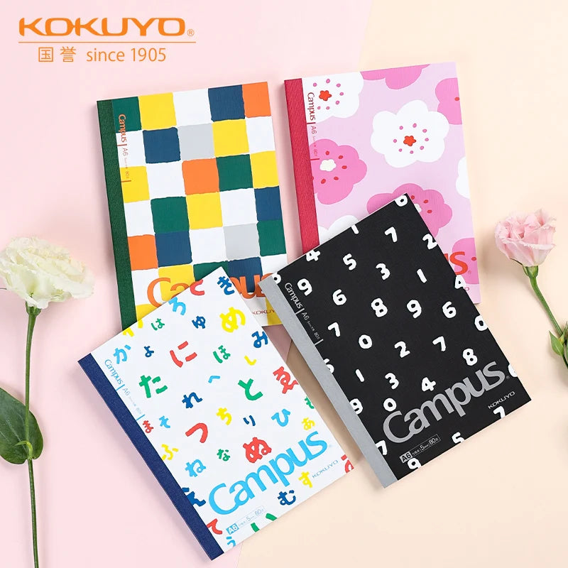 Four notebooks with different designs on a light pink background, featuring the Kokuyo brand.