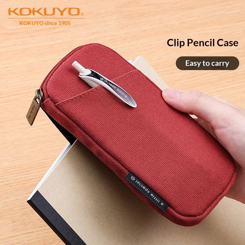 Red clip pencil case held by a hand with a pen, on a wooden surface, featuring the Kokuyo brand.