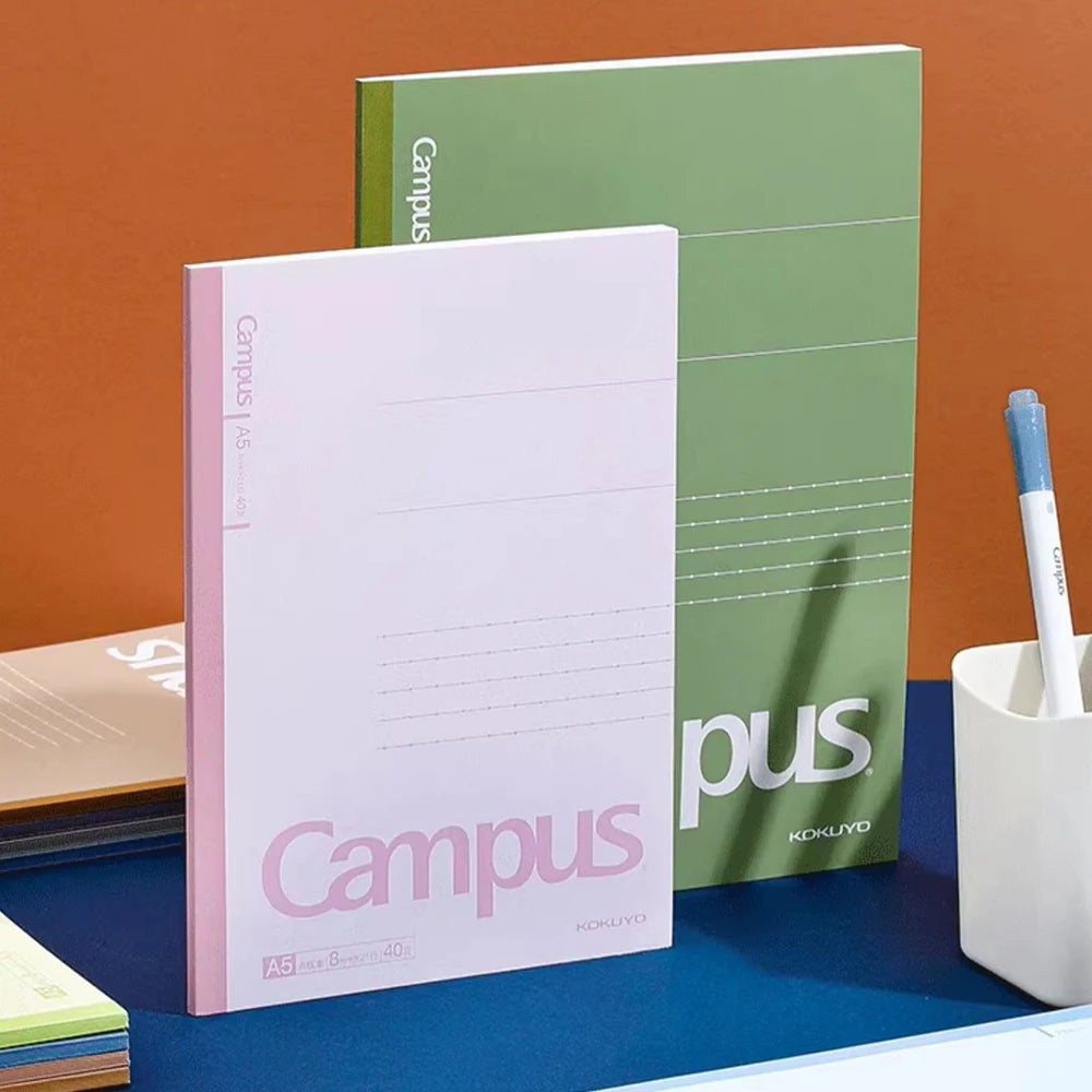 Two notebooks, one pink and one green, with 'Kokuyo' branding on a blue surface against an orange background.